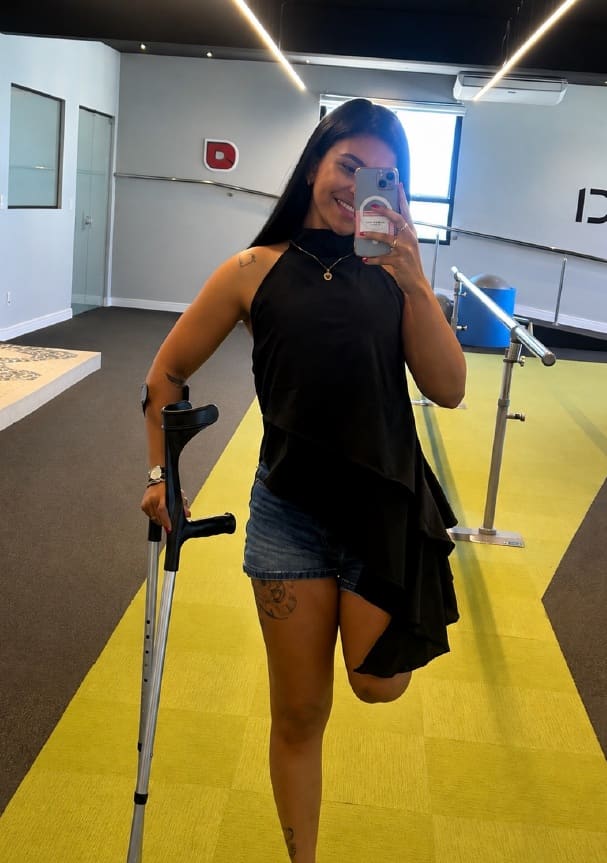 Isabella de muletas numa sala de reabilitação, fazendo selfie no espelho.