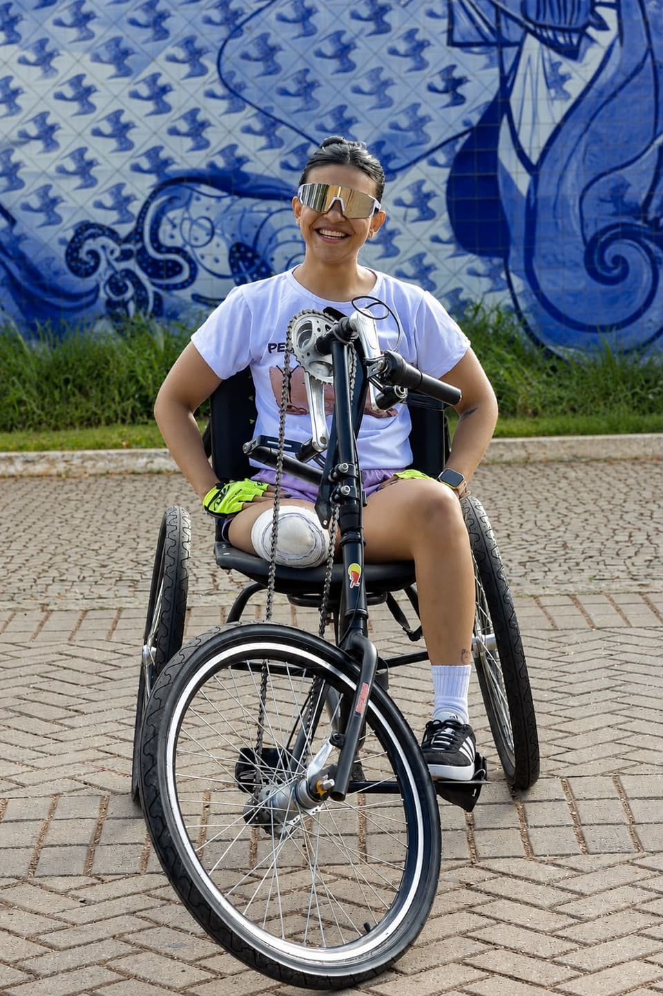 Isabella numa handbike em frente a um mural de azulejos azuis, de óculos esportivos.