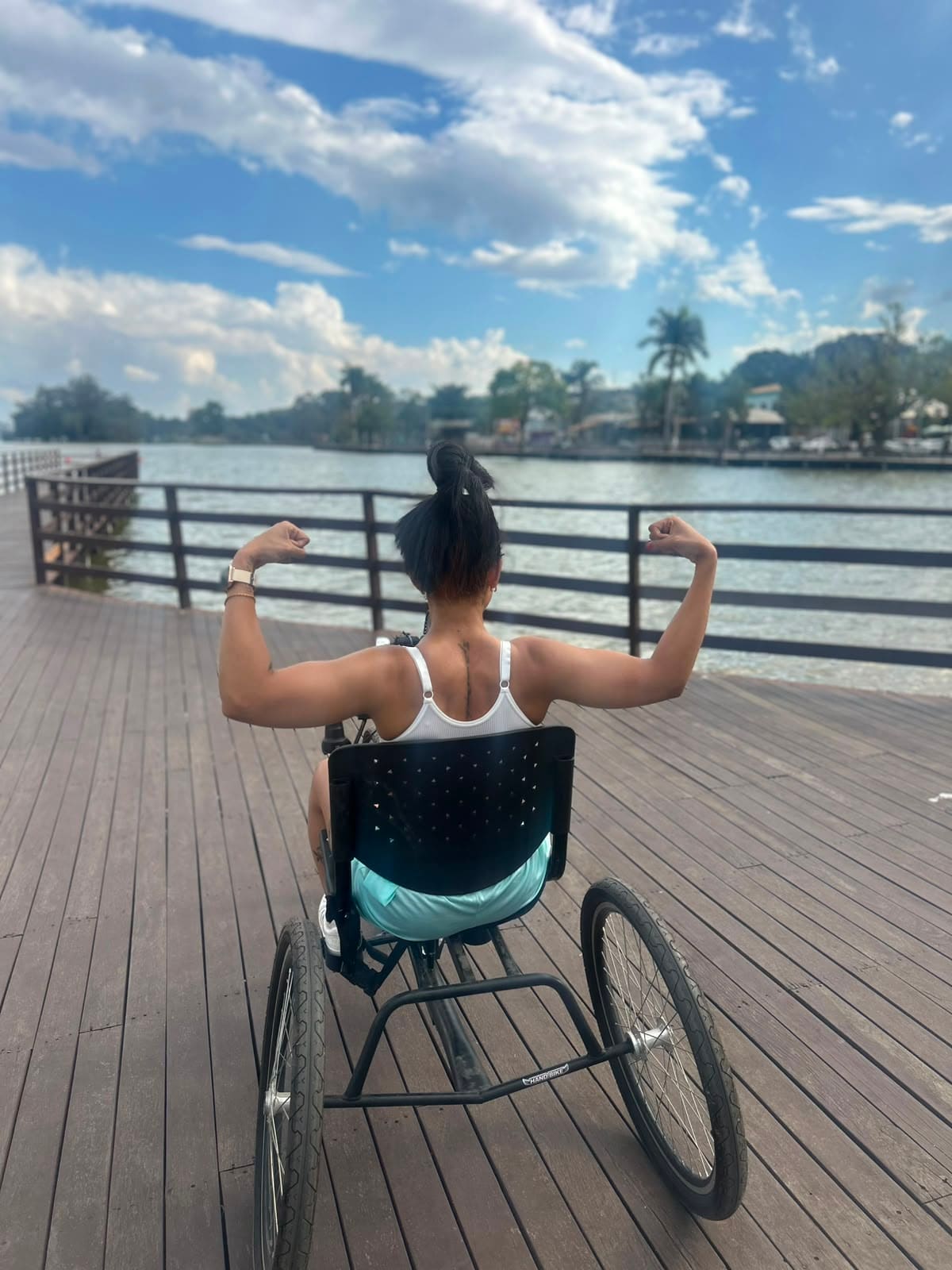 Isabella de costas numa handbike, braços erguidos, de frente para o lago.